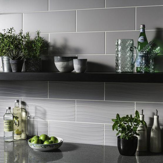 A wall with light grey satin glazed ceramic wall tiles that are rectangular and shelves with glasses, bottles and plants