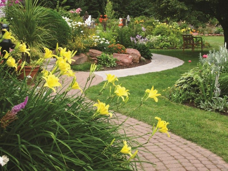 A garden with a pathway, flowers, grass, a bench and landscape fabric. 