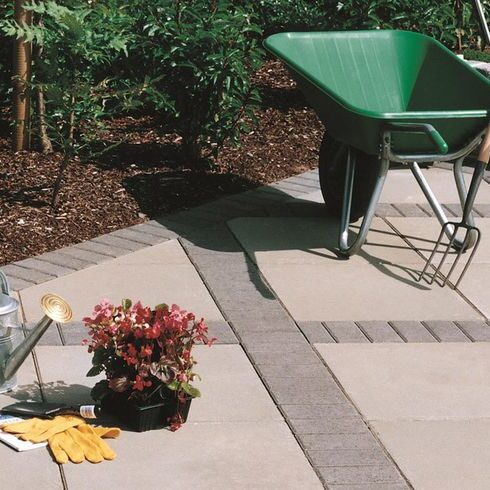 Corner of a garden paving area with a mix of beige and grey paving slabs and a wheelbarrow with a pitchfork