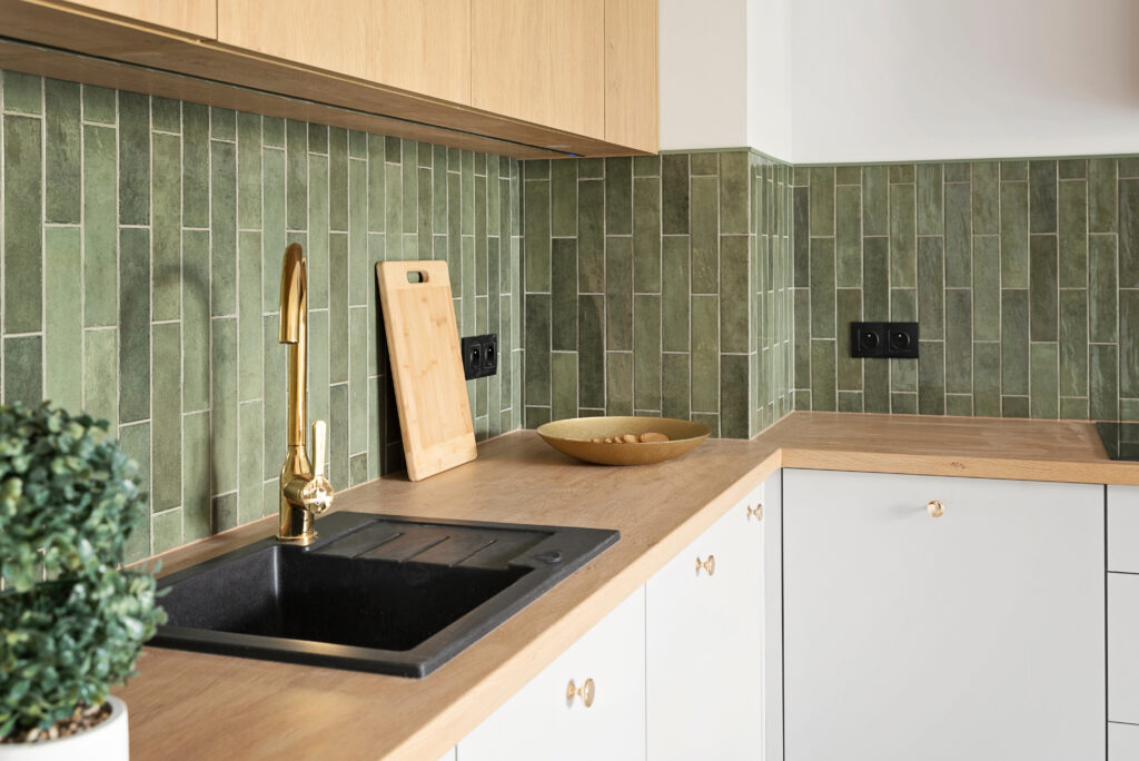 a modern kitchen with vertically placed sage green rectangular zellige tiles, light wood counter tops and white cupboards. a black sink is complemented with a gold tap