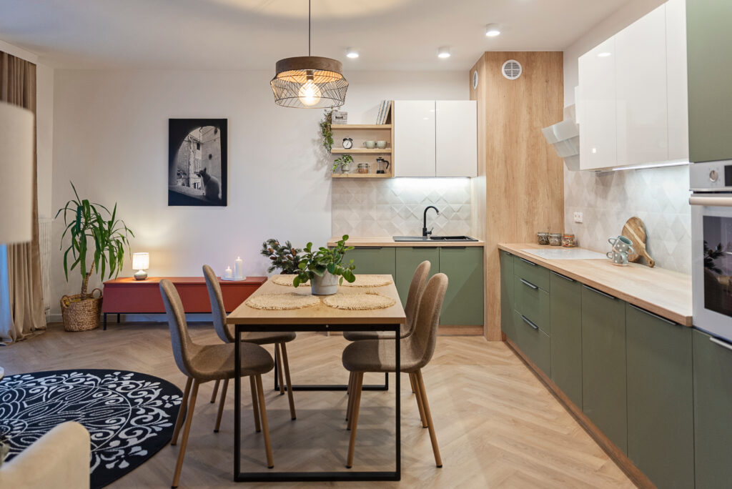 A modern kitchen with sage green cupboards, light wooden table and chairs and counter tops, with natural herringbone flooring. A black and white rug and wall art accessorise the space with warm lighting above, and a lamp sits on a red side board