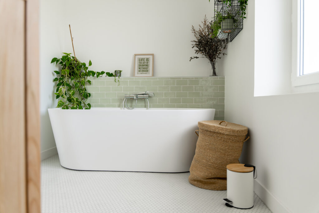 a modern white freestanding bath in a clean bathroom with a half-tiled sage green wall. Accessorised with house plants, a wicker laundry bin, and wall art 
