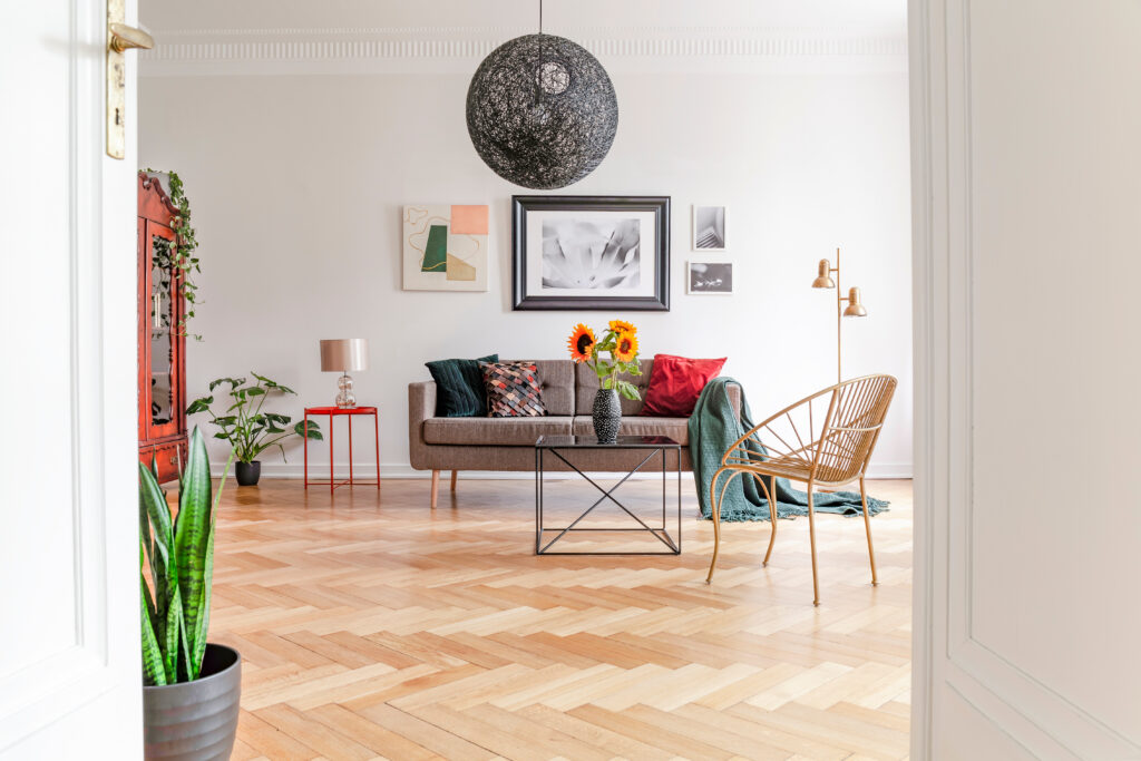 Open and airy lounge with a light coloured herringbone flooring in a zigzag pattern. The room is accessorised with a grey sofa, wicker chair, colourful cushions and wall art, a vase of sunflowers, and house plants