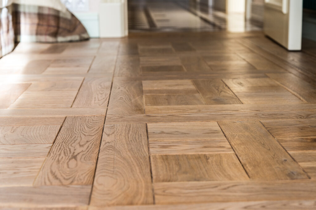 A close up of rustic wood parquet flooring with a geometric design. The blocks are arranged in a repetitive square pattern made up of rectangular edges and two shorter rectangles in the centre of each square
