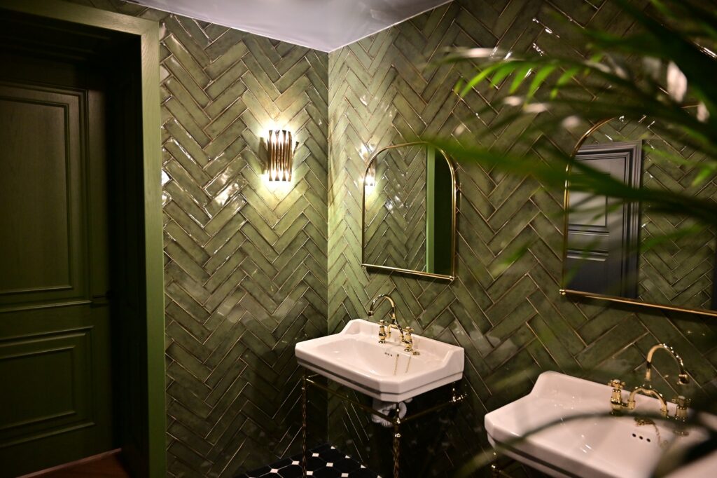 A vintage looking, olive green tiled bathroom in a herringbone style on the walls. There are two sinks side by side, with gold mirrors above them, warm lighting, and a house plant just in frame. 
