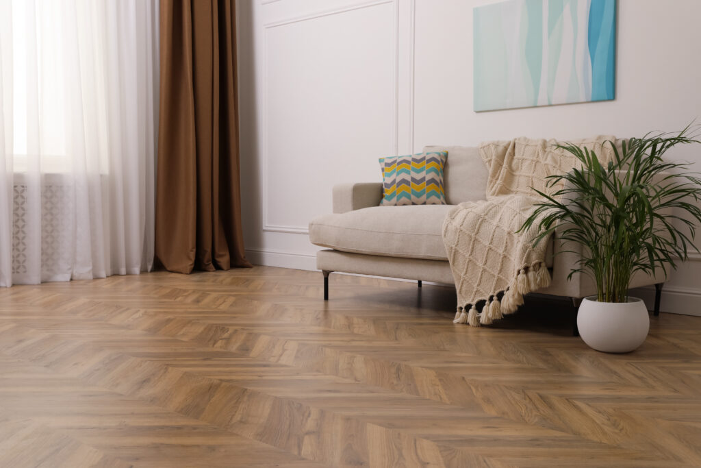 A cosy lounge with parquet chevron flooring, in a neat pattern resembling arrows. A cream sofa is placed against a white panelled wall, with a colourful zigzag cushion and cream tassel throw draped on it. Burnt orange curtains frame a window, obscured by net curtains