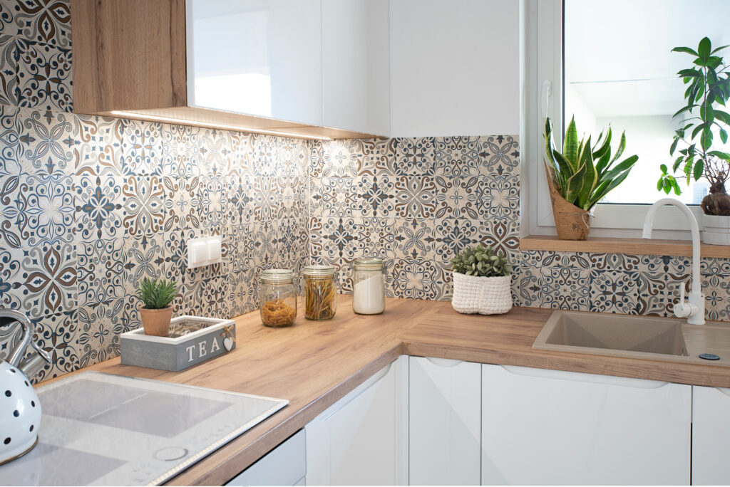 a modern kitchen with white cupboards and natural light wood counter tops features a Mediterranean inspired design on patterned wall tiles. House plants are dotted about the space