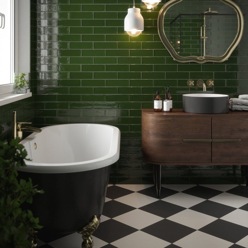 A bottle green metro tiled bathroom, with black freestanding bath by a window, dark wood vanity cabinet with sink and gold mirror above, black and white check floor tiles and gold light fixtures